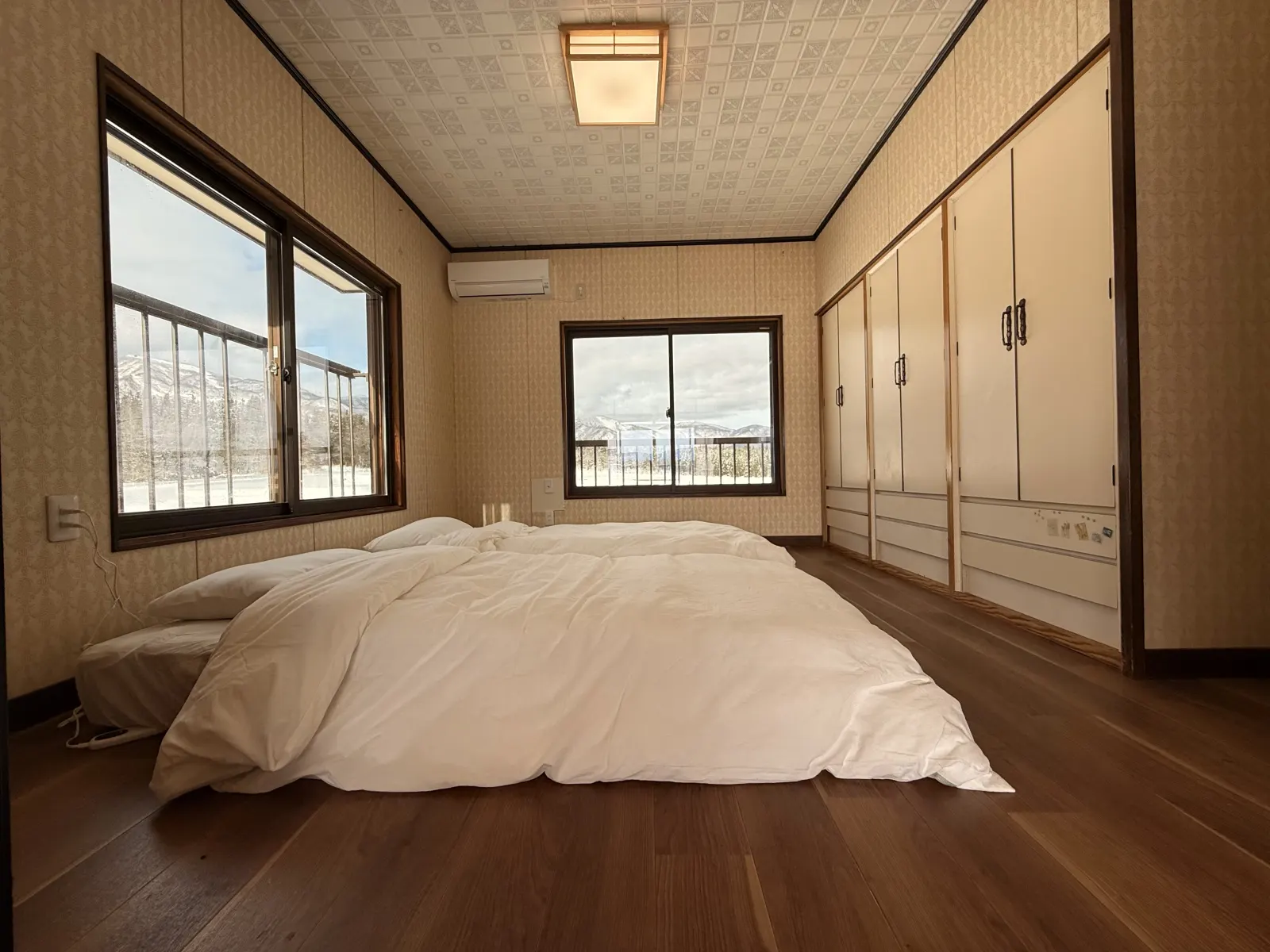 A bedroom with futons on wooden floors, built-in cupboards and two windows onto the snow and mountains.